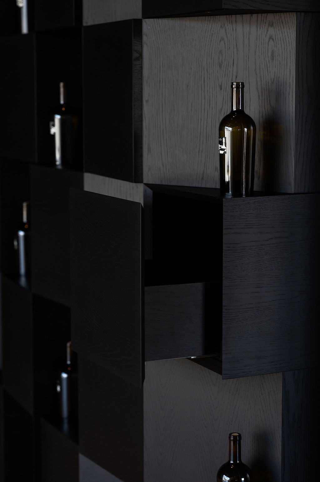 Dark wooden cabinet with a bottle inside, surrounded by similar cabinets. Checkmate Winery tasting bar and custom millwork by BWP Spaces, South Okanagan