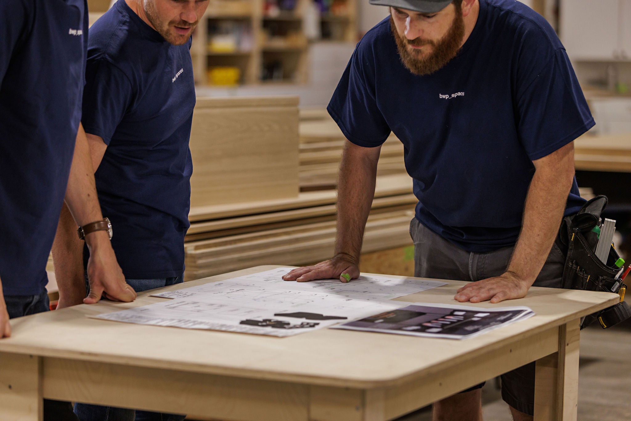 Three men in navy blue shirts discussing plans on a wooden table with wood stacks in the background.