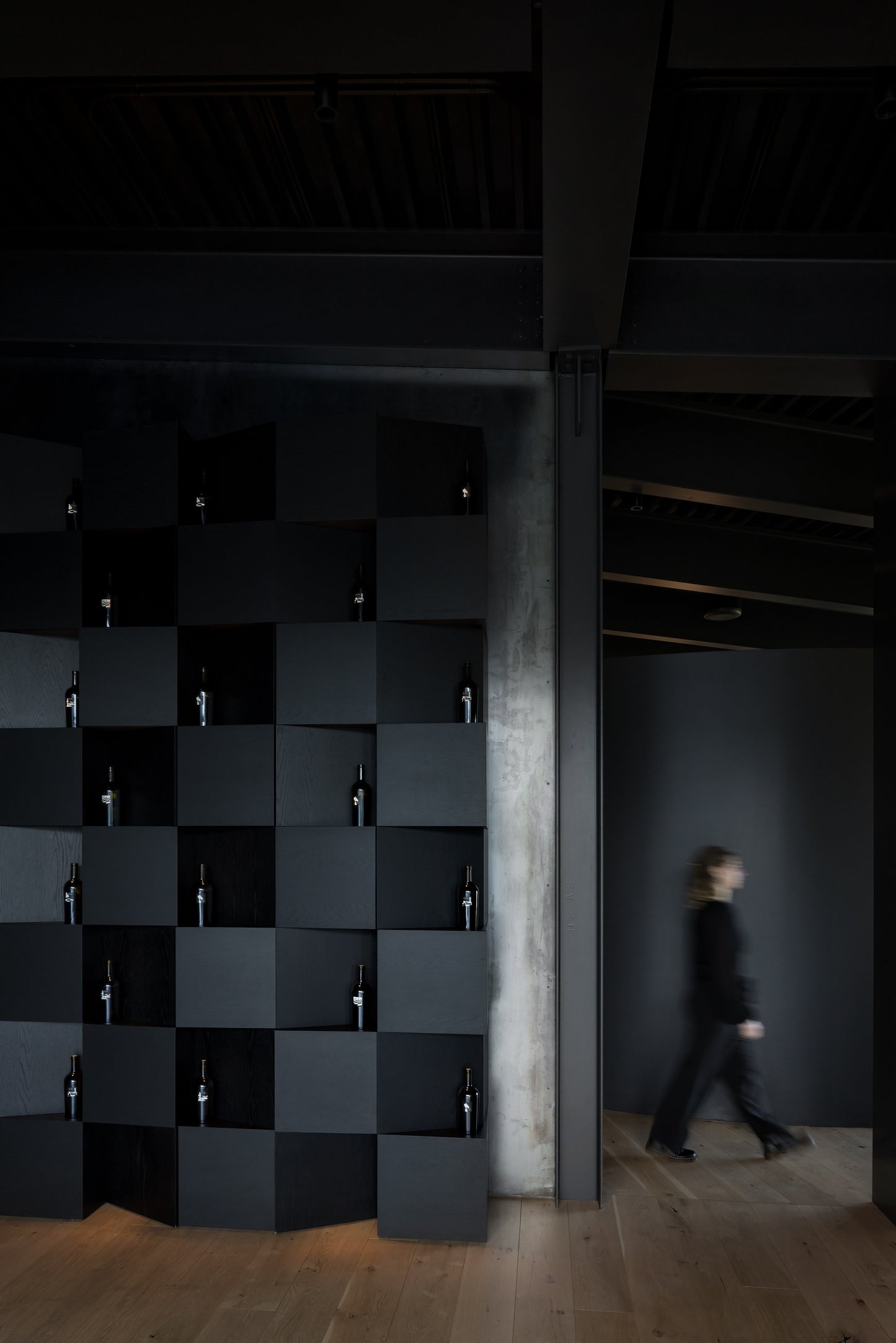A wall-mounted wine bottle display and storage unit composed of black wooden cubes, with a person walking by in the background. BWP Spaces bespoke furniture and millwork at Checkmate Winery, BC