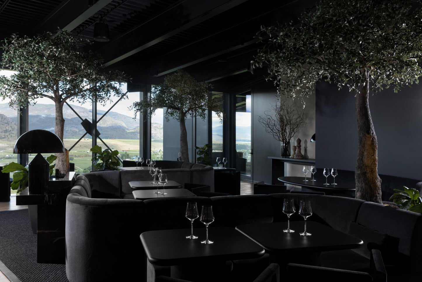 A sleek modern wine tasting lounge with dark furniture and large windows looking out onto a scenic landscape. Checkmate Winery tasting bar and custom millwork by BWP Spaces, South Okanagan